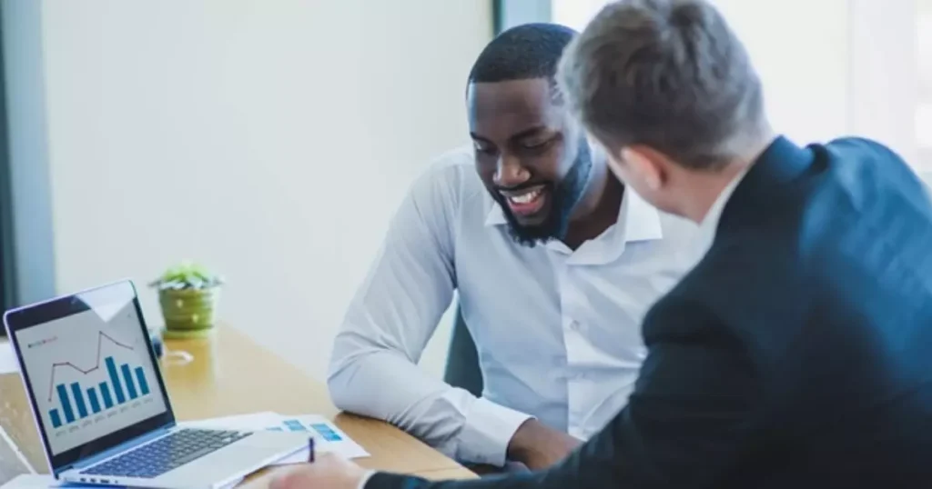 Imagem de dois homens conversando em frente de um computador, em um ambiente de trabalho, trocando um feedback em um ambiente corporativo.